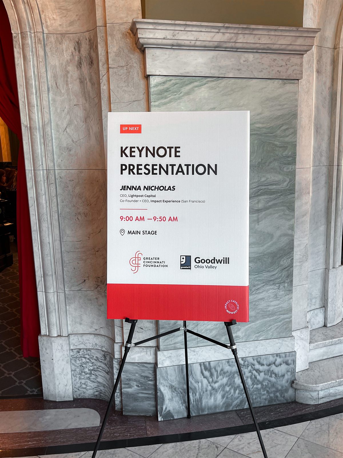 Keynote easel sign in the marble lobby listing the speaker, time, and sponsors, with the badge tucked in the red footer