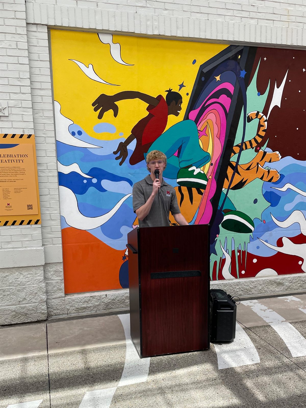 Riley speaking at a podium in front of the finished mural during the unveiling event