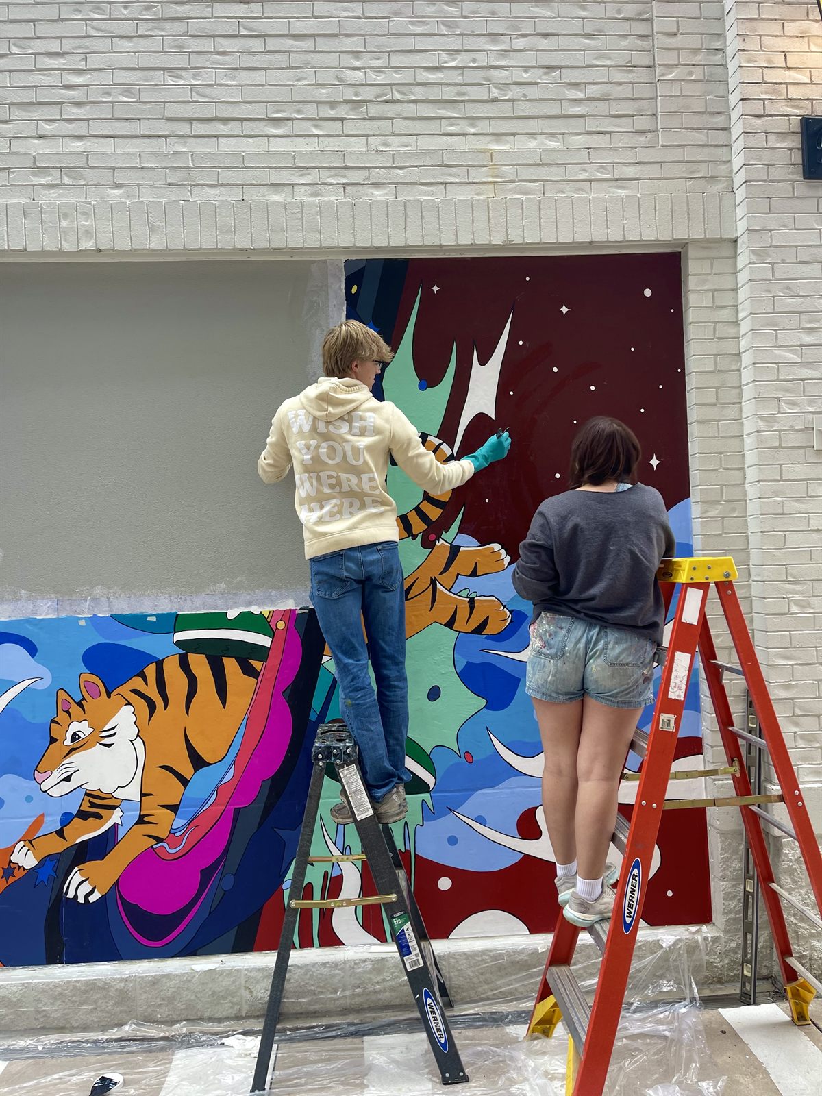 Two student artists on ladders painting mural panels of a tiger and blue waves onto the wall