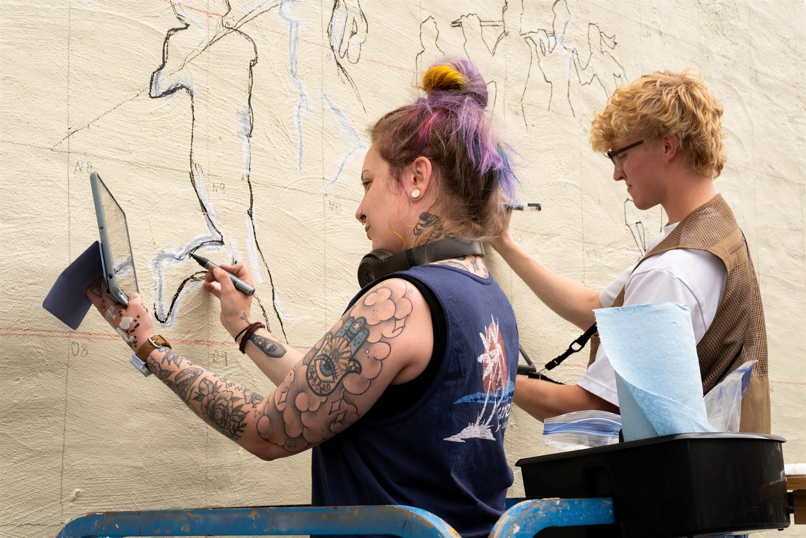 Two artists drawing the mural's outlines onto the bare wall, one working from an iPad reference